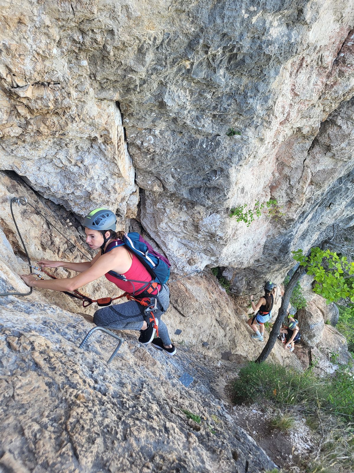 Grimpeuse casquée progressant sur le câble de la via ferrata de Liaucous, deux participants en contrebas, au-dessus des gorges du Tarn et de la Jonte près de Millau