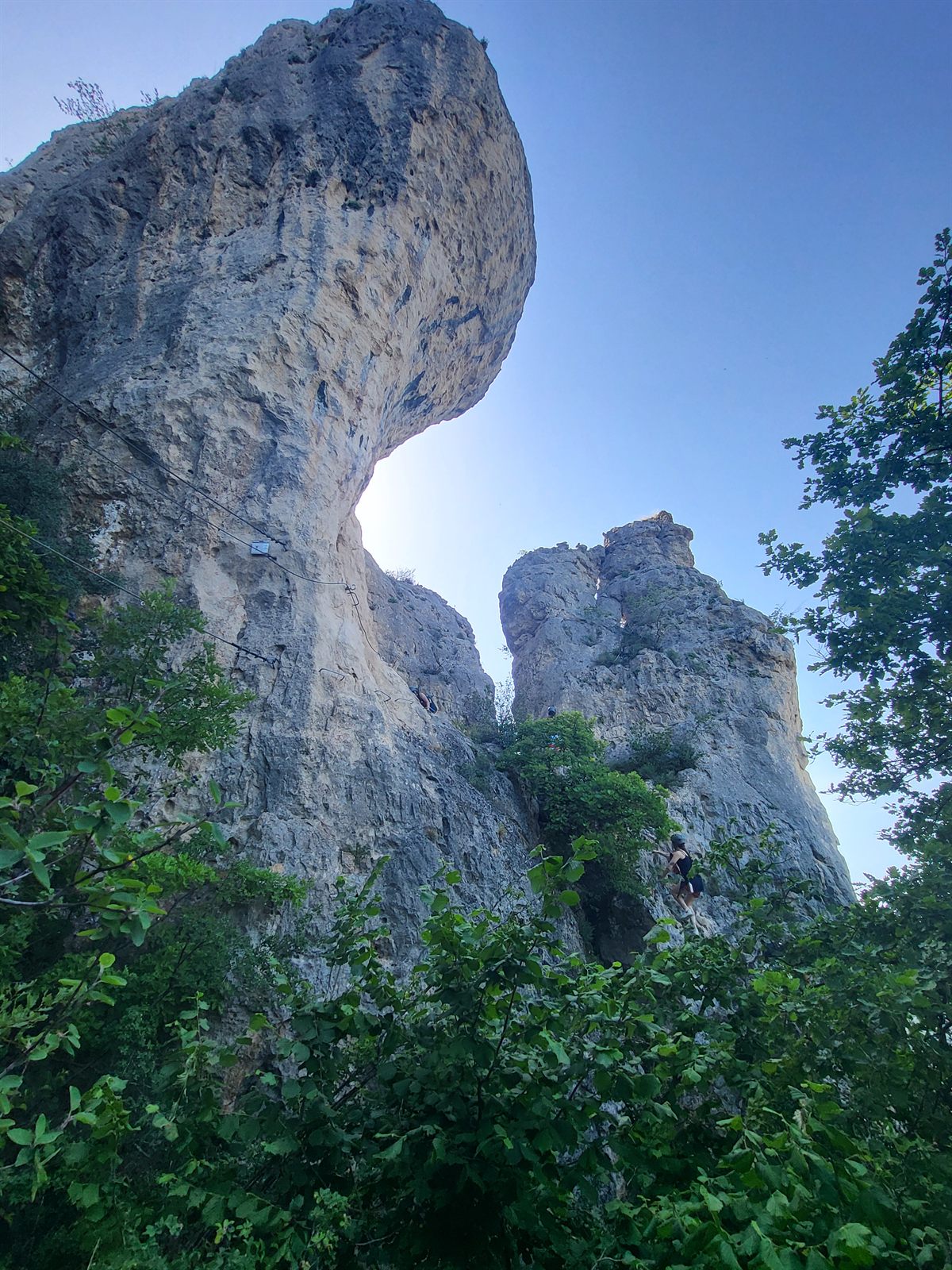 Rocher en équilibre et grand surplomb calcaire sur la via ferrata de Liaucous, ciel bleu et grimpeur au pied du pilier, gorges du Tarn et de la Jonte près de Millau