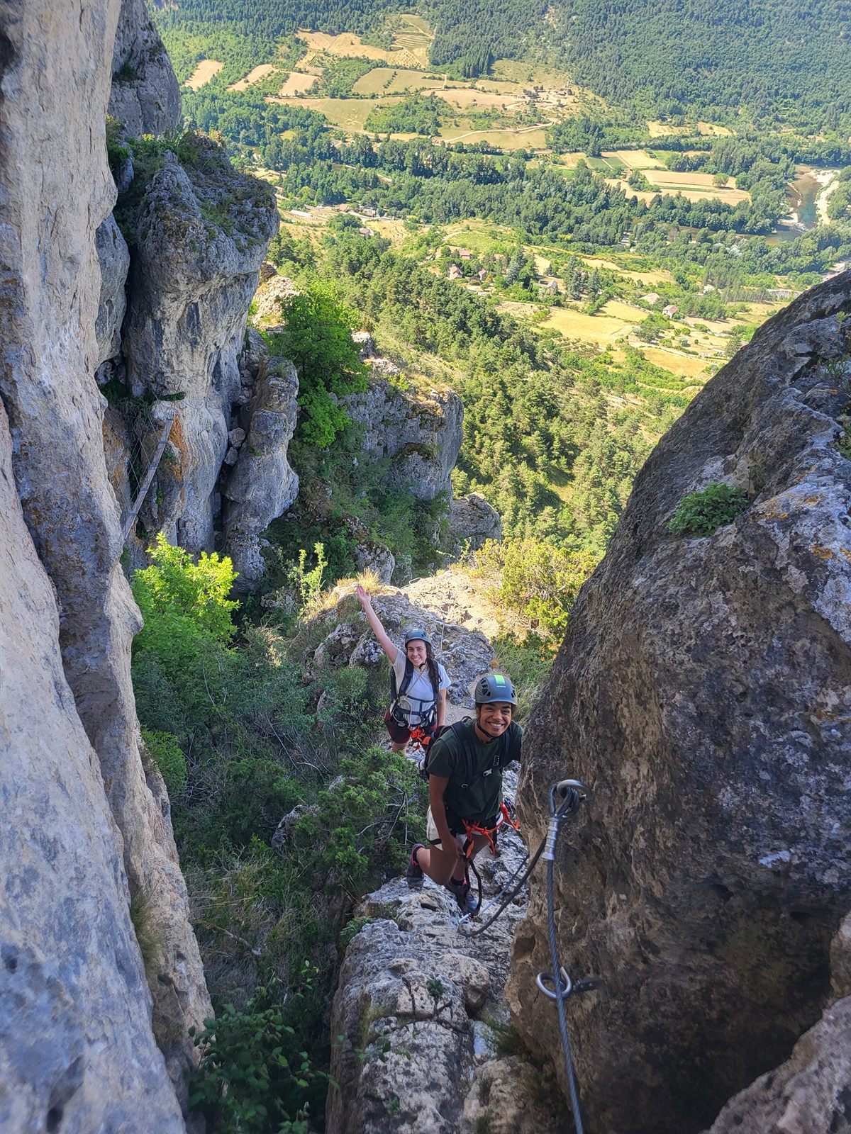 Deux participants souriants sur une vire de la via ferrata de Liaucous, panorama sur la vallée du Tarn cultivée en contrebas, près de Millau
